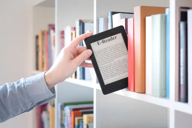 Dłoń wyjmująca czytnik e-booków z półki między książkami
