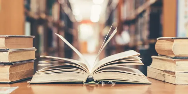 Otwarta książka na stosie w bibliotece, w tle rozmyte regały